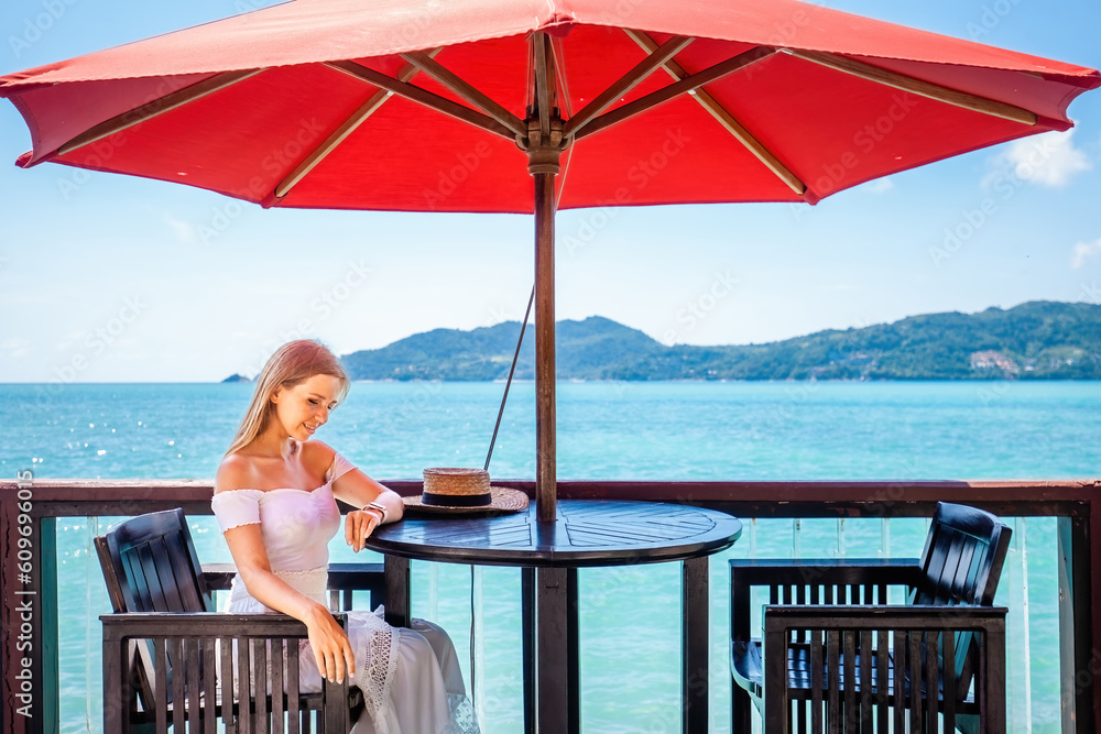 Young woman enjoys a romantic dinner at a restaurant on the beach. Girl sitting at a table by the water, enjoying the sound of the waves. Vacation, summer, travel, holidays concept