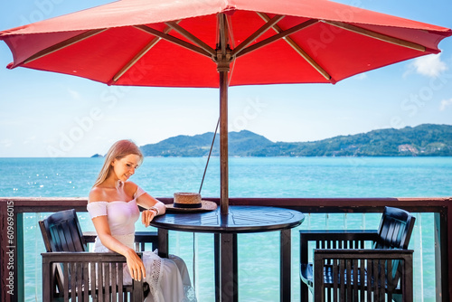 Young woman enjoys a romantic dinner at a restaurant on the beach. Girl sitting at a table by the water, enjoying the sound of the waves. Vacation, summer, travel, holidays concept © TravelMedia