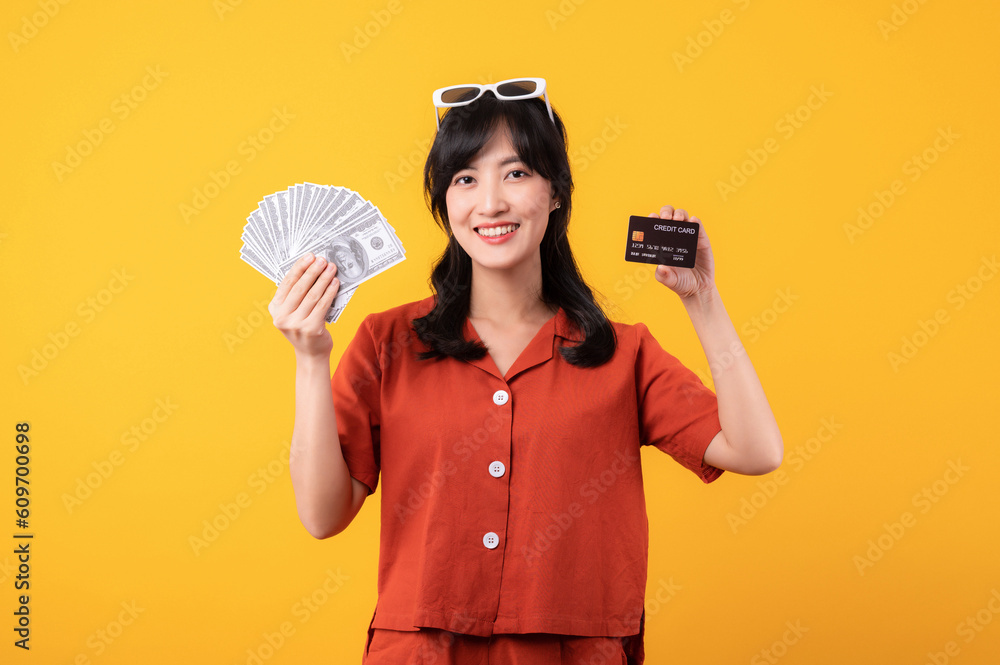 portrait of happy successful confident young asian woman happy smile wearing orange clothes holding cash money and credit card standing over yellow background. millionaire business, shopping concept portrait of happy successful confident young asian woman happy smile wearing orange clothes holding cash money and credit card standing over yellow background. millionaire business, shopping concept
