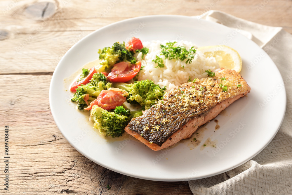 Crispy fried salmon filet with broccoli tomato vegetable and rice, parsley and lemon zest garnish, served on a white plate on a rustic wooden table, copy space, selected focus