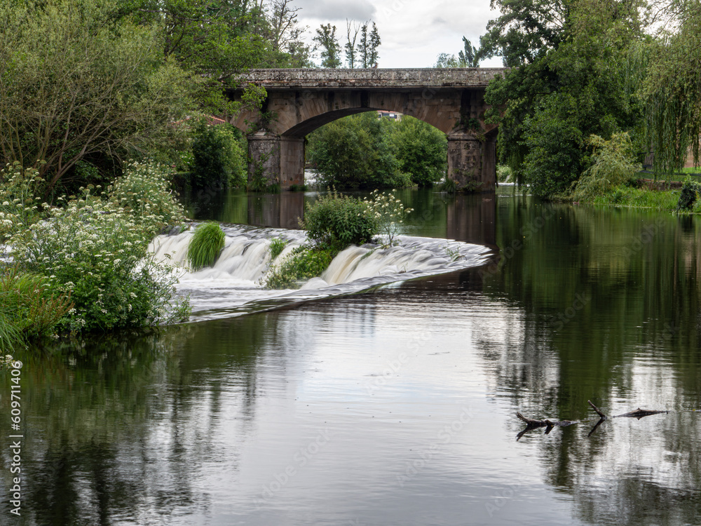 View of the bridge reflected on the river that surrounds the green nature and a small waterfall on a bank with a curve shape, in Allaritz, Galicia, summer 2021 View of the bridge reflected on the river that surrounds the green nature and a small waterfall on a bank with a curve shape, in Allaritz, Galicia, summer 2021