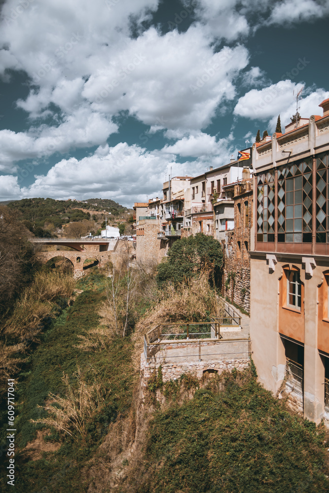 Caldes de Montbui; a vertical shot of the blue sky with clouds. Lush vegetation is located behind the warm-colored buildings, adding a natural backdrop. A charming Roman bridge completes the scene