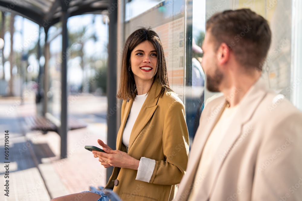 Happy stylish modern young woman talking to unrecognizable man in beige suit while waiting for bus and sitting at bus station in the city. Happy stylish modern young woman talking to unrecognizable man in beige suit while waiting for bus and sitting at bus station in the city.