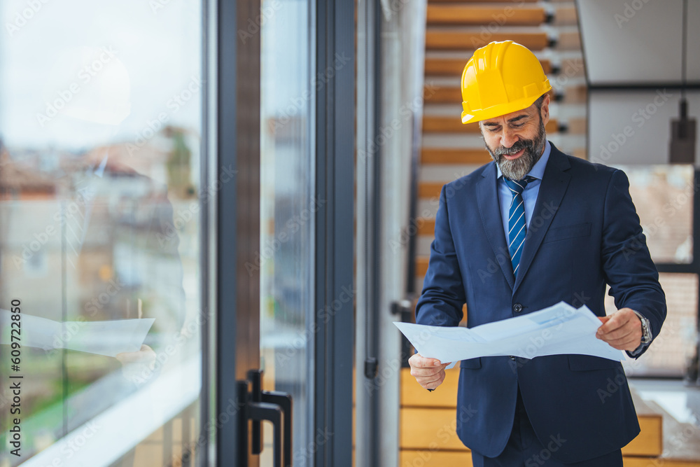 Portrait of an engineer holding a blueprint at a construction site while wearing a helmet. Construction worker with helmet on construction site examining office blueprints. Indoors Portrait of an engineer holding a blueprint at a construction site while wearing a helmet. Construction worker with helmet on construction site examining office blueprints. Indoors