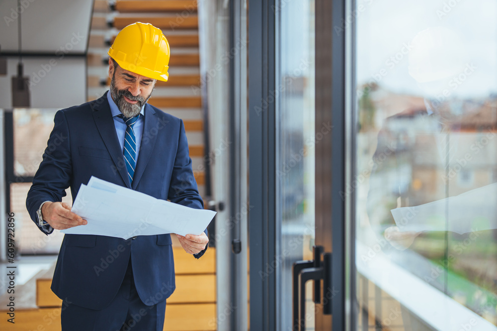 Construction engineer or architect with blueprints visiting and chechking eco building site. Smart architect examining draft during work. Mature man in a suit and construction hat Construction engineer or architect with blueprints visiting and chechking eco building site. Smart architect examining draft during work. Mature man in a suit and construction hat
