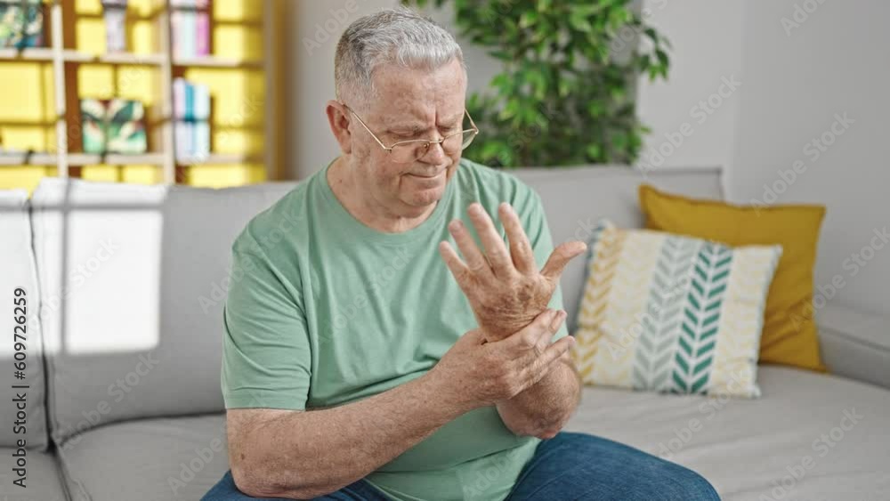 Middle age grey-haired man sitting on sofa suffering for wrist pain at home Middle age grey-haired man sitting on sofa suffering for wrist pain at home