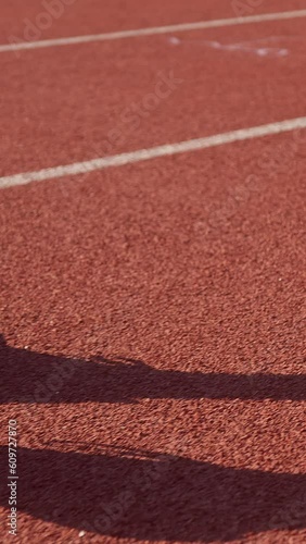 Vertical video of male Caucasian athlete legs in black sneakers starting to run, striding on the red athletic track, slider shot.