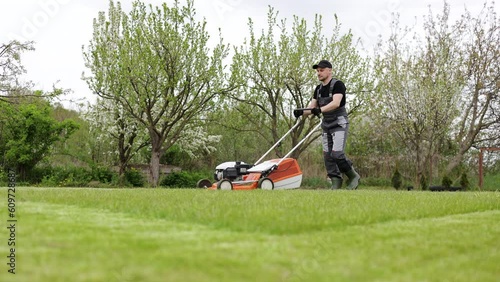 Professional gardener in protective apparel is mowing green grass lawn using modern gasoline cordless lawnmower at backyard. Seasonal landscaping design work. Blooming trees on background.