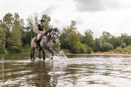 Girl riding gray horse down the calm river water with forest greenery reflections from the background. Nature and animal love concept.