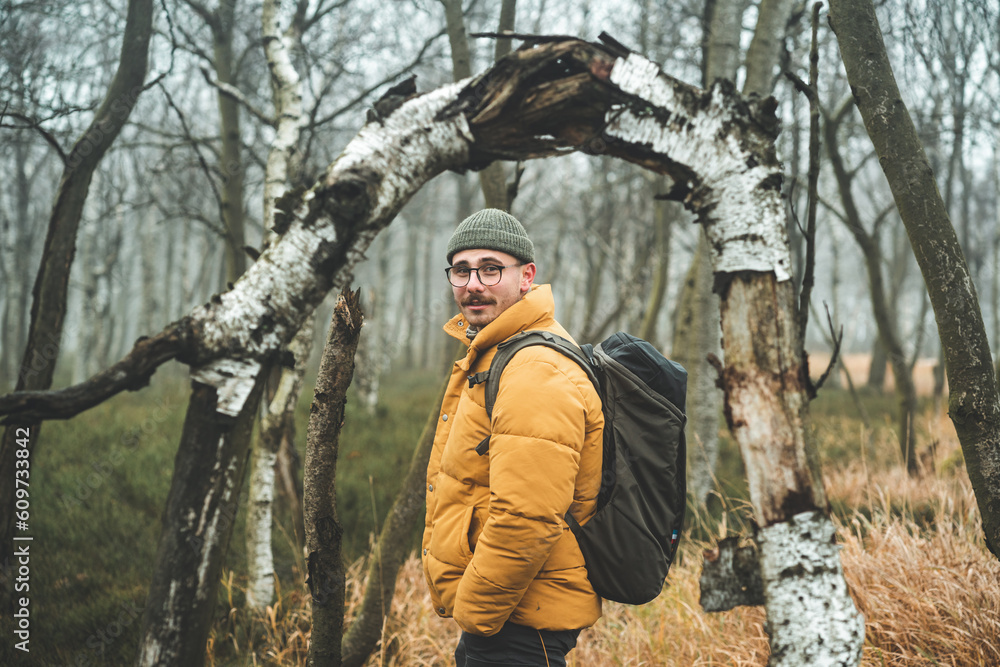 Portrait of young smiling friendly man with yellow winter jacket and hat, that looks behind his shoulder and stands under bending tree, during cold cloudy autumn day, Decin, Czech republic Portrait of young smiling friendly man with yellow winter jacket and hat, that looks behind his shoulder and stands under bending tree, during cold cloudy autumn day, Decin, Czech republic