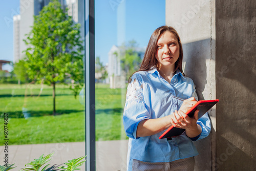 Serious confident office worker holding tablet by window. Caucasian young woman. Business
