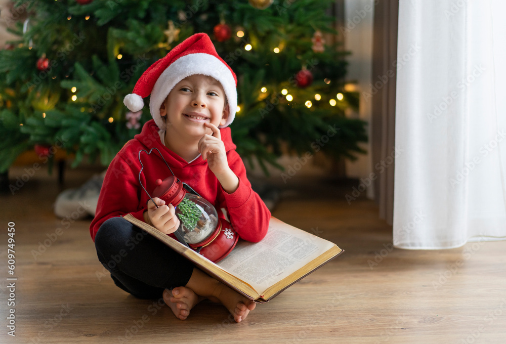 A little boy in a Santa Claus hat sits on the floor and reads a book with fairy tales A little boy in a Santa Claus hat sits on the floor and reads a book with fairy tales
