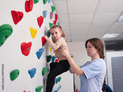 physiotherapist instructor helping little girl to climb wall in gym. sensory integration for kid and correctional physiotherapy to replenish sensory experience