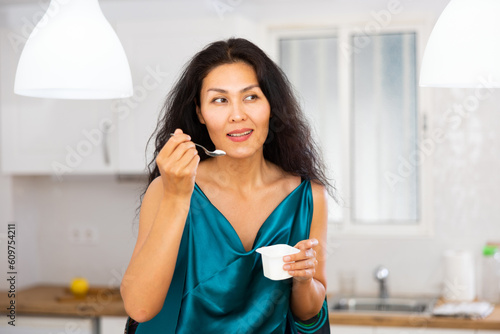 Portrait of asian woman in nightie eating yoghurt in kitchen at home.