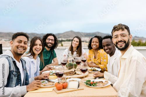 Group of friends gathered smiling at camera sitting together at a table enjoying barbecue dinner celebrating a party in summer. Portrait of multiracial people