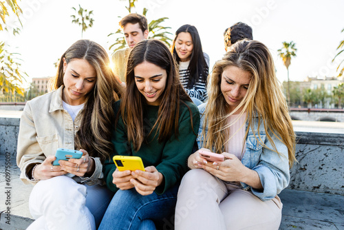 Teenage group of young people looking at smart mobile phone screen outdoors. Addicted millennial student friends using smartphones to watch social media content sitting together in city street.