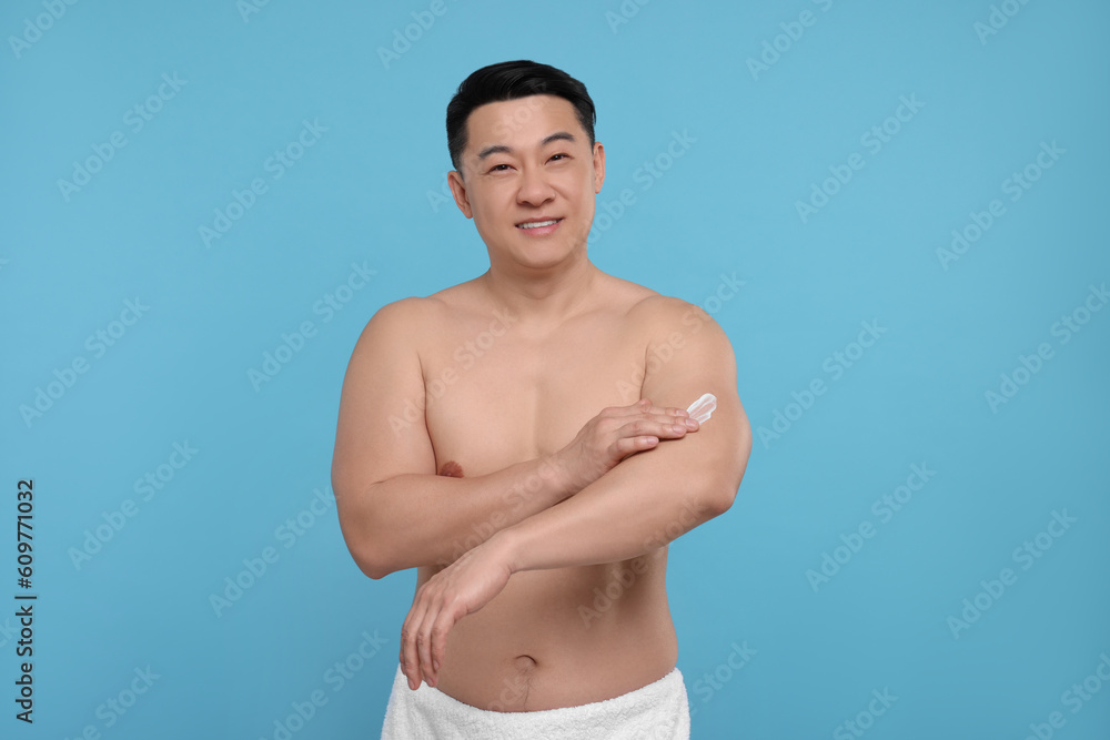 Handsome man applying body cream onto his arm on light blue background
