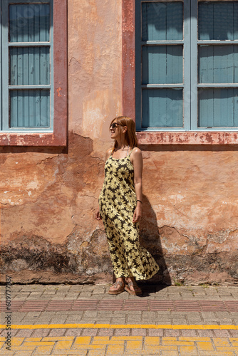 ginger woman standing posing relaxed looking at horizon in yellow and black flowery dress on pink facade background of picturesque house