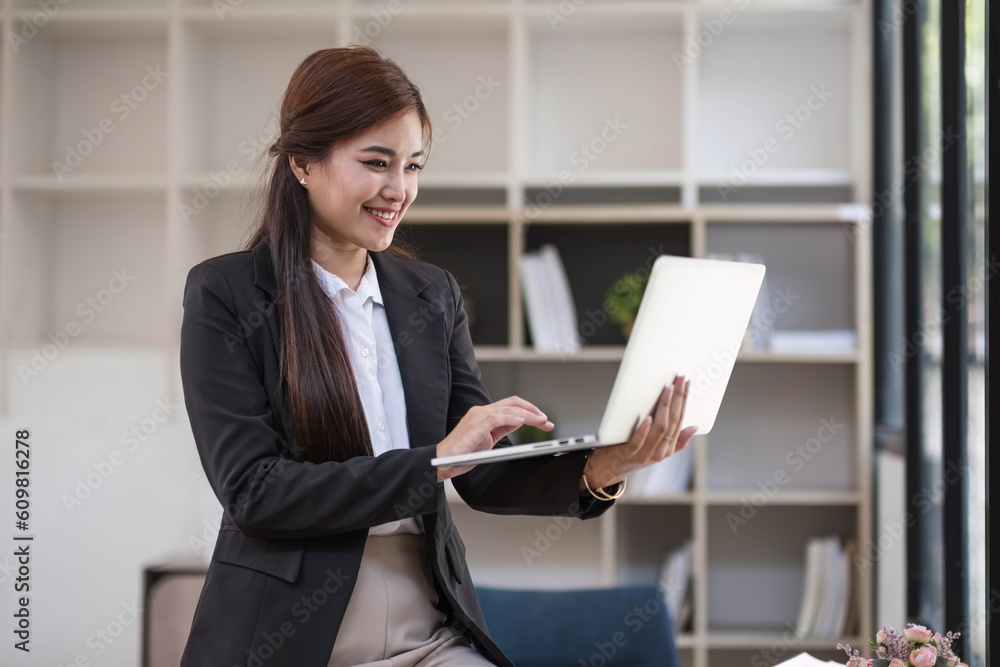 Portrait, Professional and confident millennial Asian businesswoman or female executive manager in formal suit and eyeglasses, standing, leaning on table, holding laptop and using laptop computer. Portrait, Professional and confident millennial Asian businesswoman or female executive manager in formal suit and eyeglasses, standing, leaning on table, holding laptop and using laptop computer.