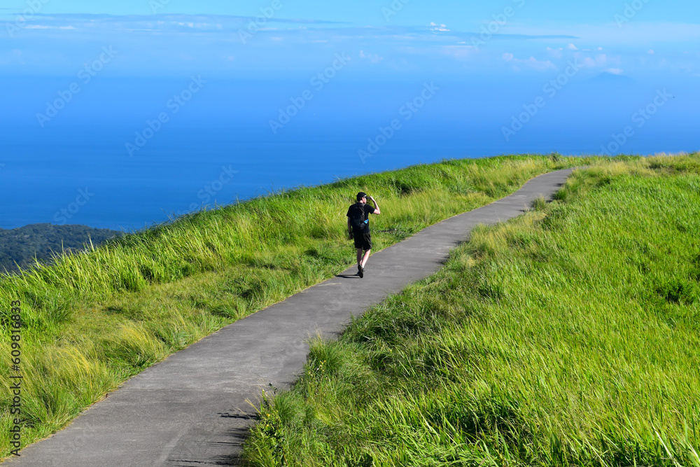 青いそら青い海。緑の山道を歩くバックパックを背負っている男性。