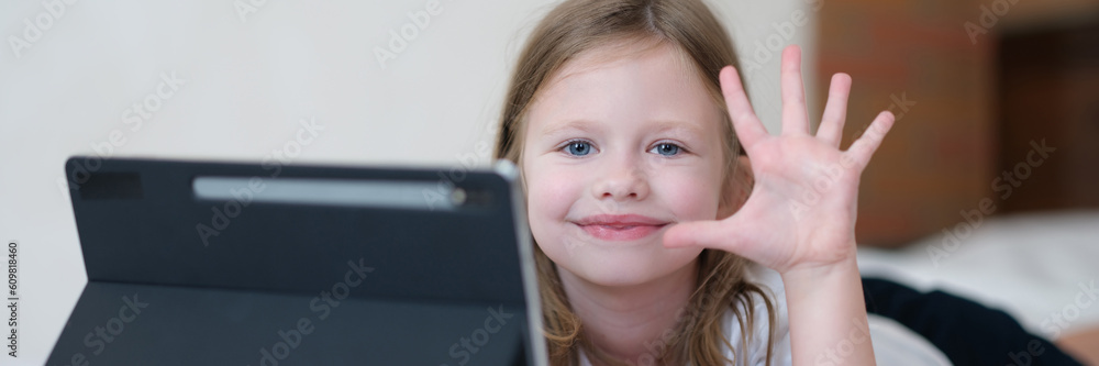 Portrait of a smiling child girl with tablet on bed holds hand in greeting. Gadgets for development and games in children Portrait of a smiling child girl with tablet on bed holds hand in greeting. Gadgets for development and games in children