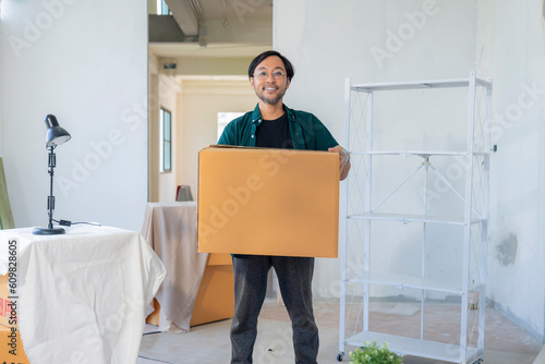 home moving relocation smiling cheerful Asian male with glasses standing hand hold cardboard home stuff standing in new home apartment look at camera,new home renovation house owner portrait with box