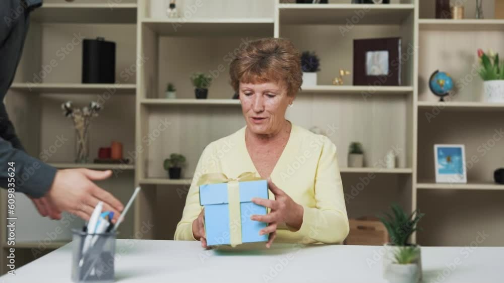 An elderly woman is sitting at a table, and she is handed a gift in a blue box with a golden ribbon. The woman is happy and smiling as she opens it. An elderly woman is sitting at a table, and she is handed a gift in a blue box with a golden ribbon. The woman is happy and smiling as she opens it.
