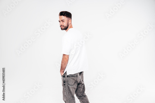 Portrait of handsome smiling stylish hipster lambersexual model. Sexy man dressed in white T-shirt and jeans. Fashion male isolated on white background in studio. Turns around