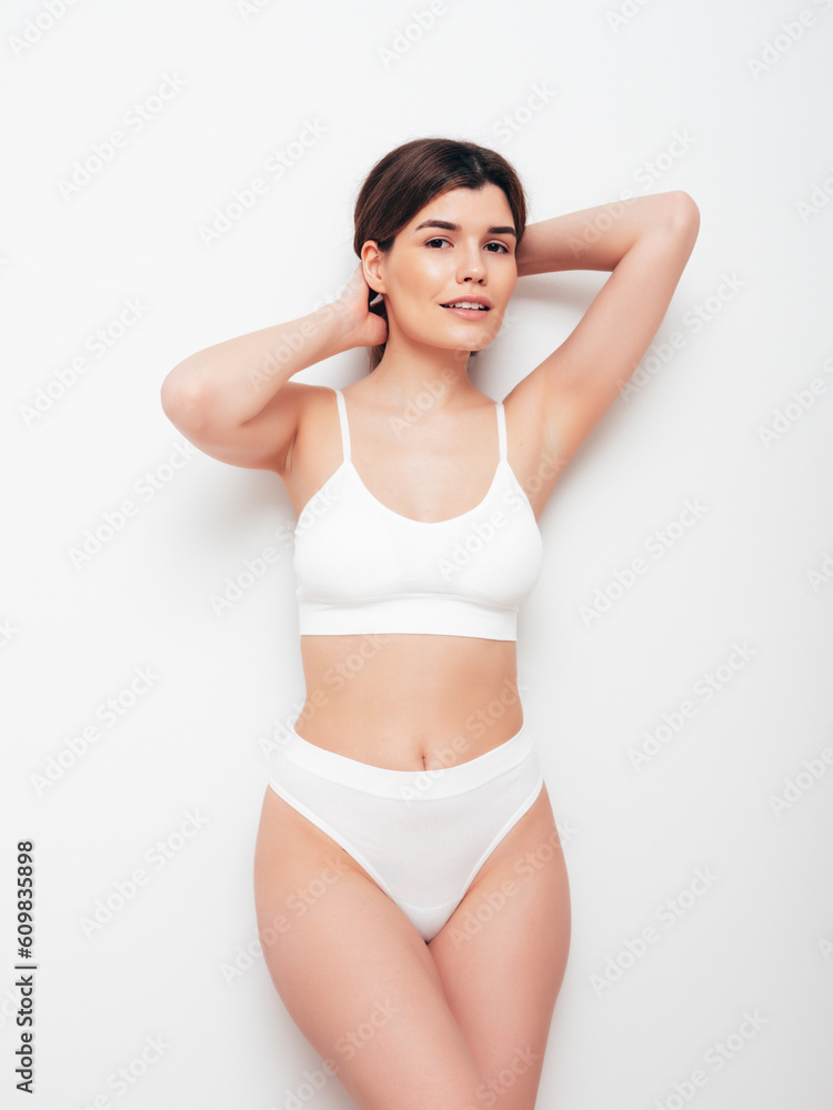 Obraz premium Portrait of young beautiful sexy woman in white lingerie. Smiling carefree model wearing pure underwear. Hot brunette posing near wall in studio interior. Perfect body. Slim and fit