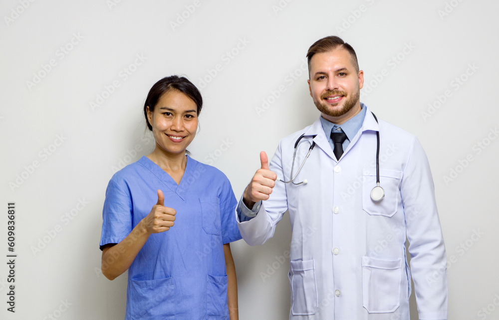 Young caucasian doctor and asian physical therapist stand with finger thumb up in front of white wall. Young caucasian doctor and asian physical therapist stand with finger thumb up in front of white wall.