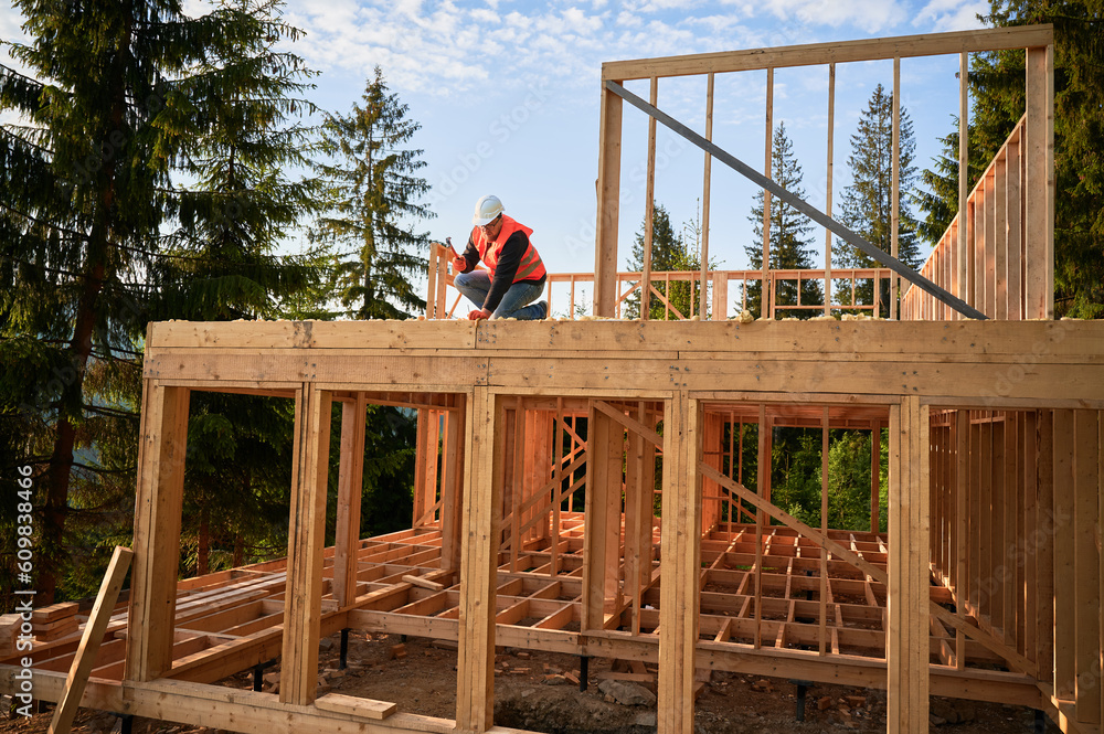 Carpenter constructing two-story wooden frame house near forest. Bearded man hammering nails into the structure, wearing protective helmet, construction vest. Concept of modern ecological construction Carpenter constructing two-story wooden frame house near forest. Bearded man hammering nails into the structure, wearing protective helmet, construction vest. Concept of modern ecological construction