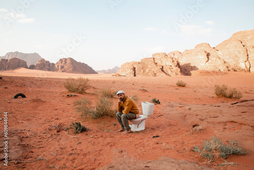 Thoughtful man sitting on toilet seat and defecating in desert photo