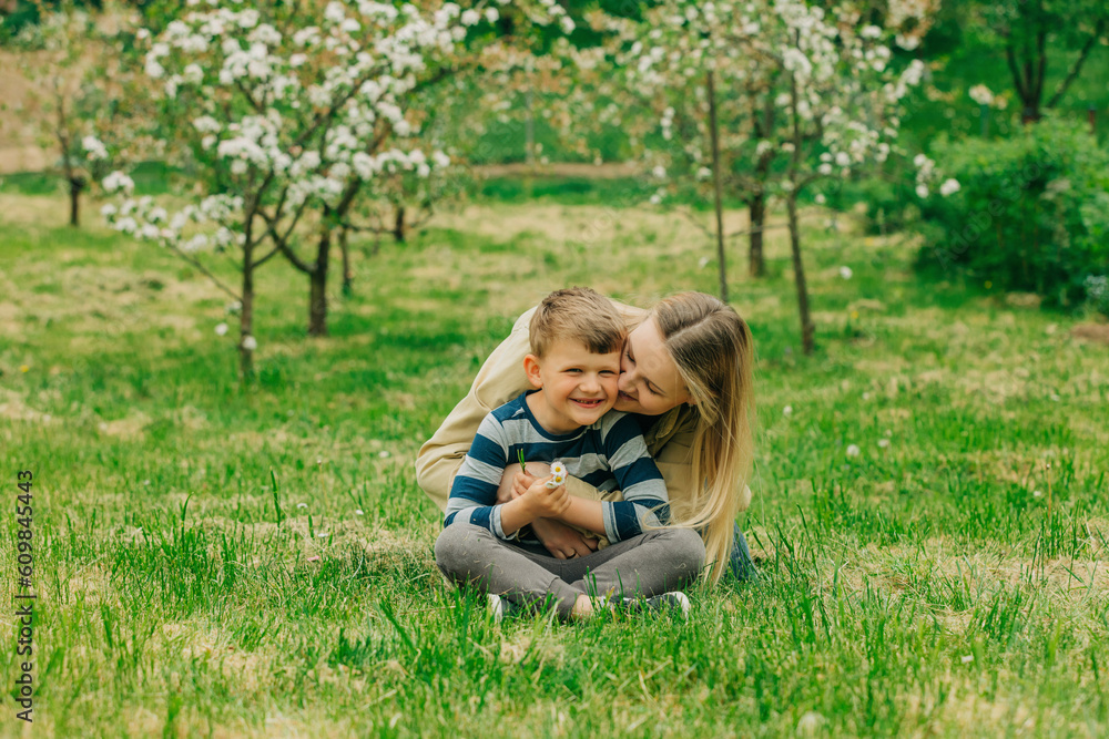 Happy mother hugging and kissing son sitting on grass in garden Happy mother hugging and kissing son sitting on grass in garden