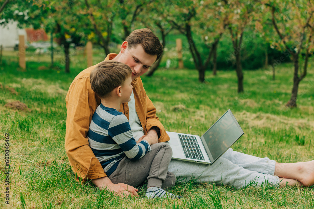 Son and father sitting with laptop on grass in garden Son and father sitting with laptop on grass in garden