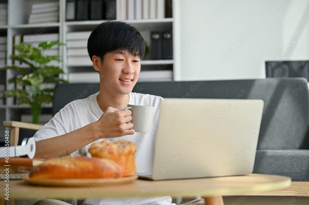 A happy young Asian man is sipping coffee while reading something on a website on his laptop