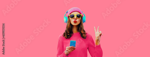 Trendy colorful portrait of stylish young woman in headphones listening to music with phone and blowing her lips sends air kiss on pink background