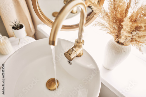 A beautiful sink with water turned on with a golden faucet next to an oval mirror and a shelf with hand towels. Close-up of an elegant golden faucet in the bathroom sink next to stylish decorations.
