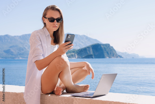 A young woman is sitting on the beach and working on a laptop while typing on a mobile phone. Remote freelance work