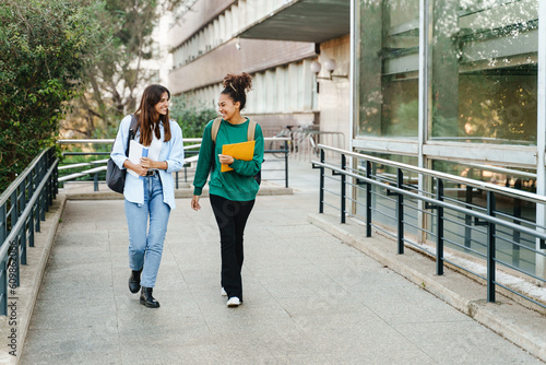 Two happy students girls walking and talking each other in University campus after classes