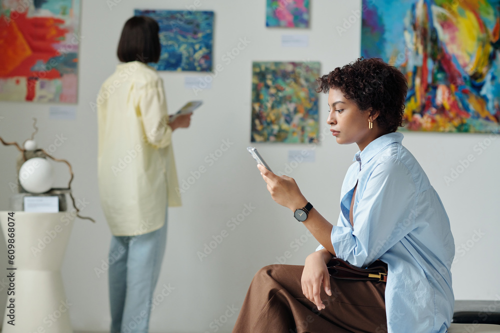 Obraz premium Young woman browsing her smartphone while resting after exhibition in art gallery