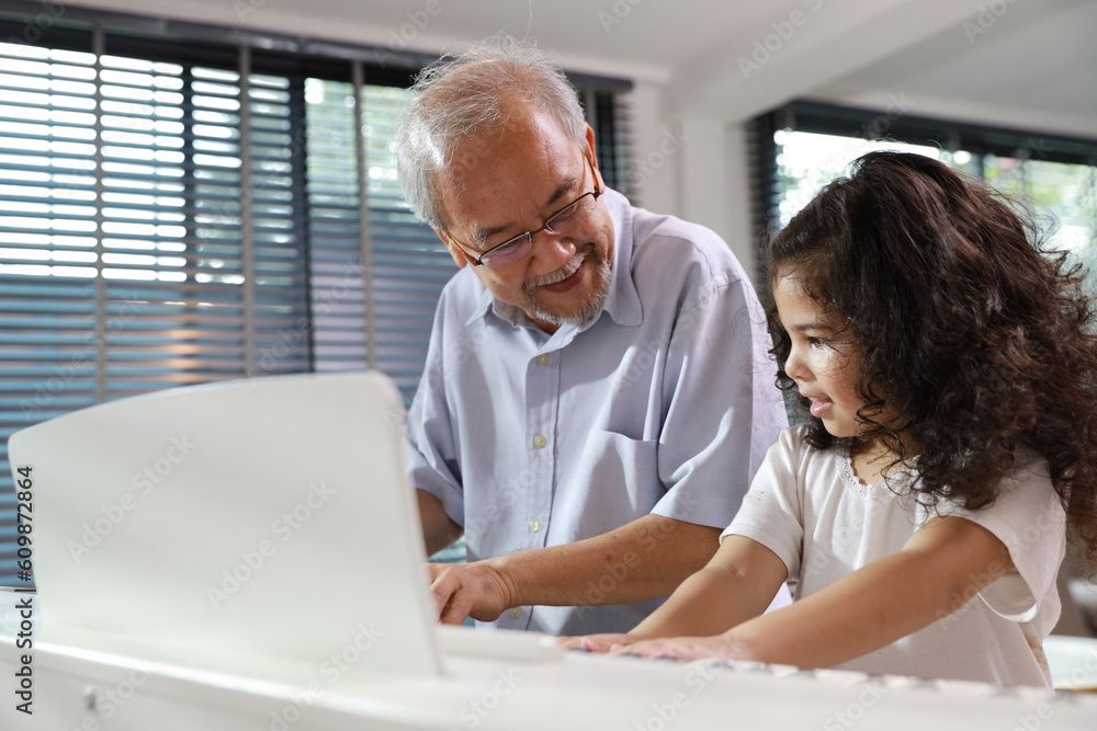 Happy smiling asian senior man sitting and playing piano while teaching grandchild in living room house indorrs. Musical and relaxation makes elder male happiness. Health care lifestyle concept. Happy smiling asian senior man sitting and playing piano while teaching grandchild in living room house indorrs. Musical and relaxation makes elder male happiness. Health care lifestyle concept.