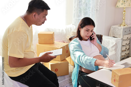 Asian couple working on box packaging, attractive young asian pregnant woman sitting and using mobile smart phone while writing order from customer with computer and husband working beside © feeling lucky
