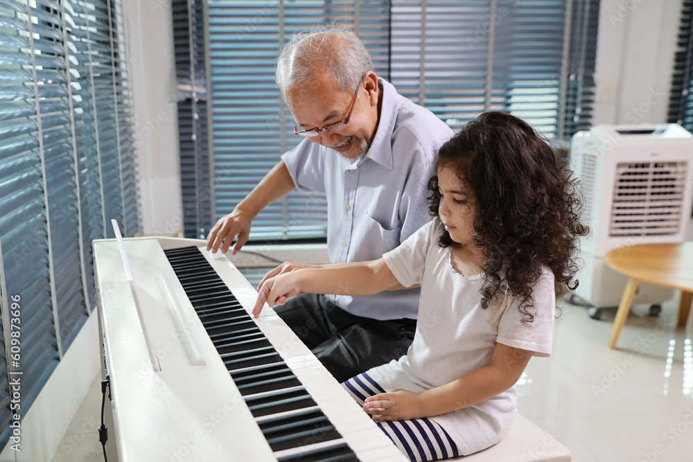 Happy smiling asian senior man sitting and playing piano while teaching grandchild in living room house indorrs. Musical and relaxation makes elder male happiness. Health care lifestyle concept. Happy smiling asian senior man sitting and playing piano while teaching grandchild in living room house indorrs. Musical and relaxation makes elder male happiness. Health care lifestyle concept.
