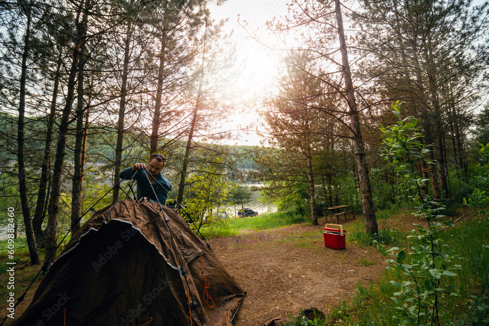 A man sets up a brown tourist tent alone. Coniferous forest, in the background an off-road vehicle, recreation and fishing on the banks of the Dniester River, Ukraine. A man sets up a brown tourist tent alone. Coniferous forest, in the background an off-road vehicle, recreation and fishing on the banks of the Dniester River, Ukraine.