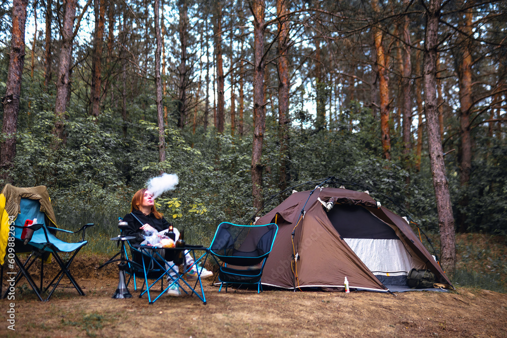 A woman exhales smoke have hookah, sits near folding table alone, drink drinks, have satisfied expressions. Pine forest, summer day. Travel and vacation. Camping tent.