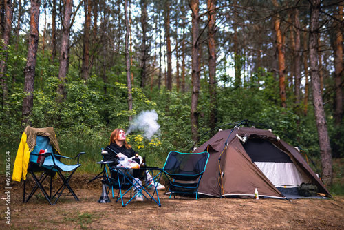 A woman exhales smoke have hookah, sits near folding table alone, drink drinks, have satisfied expressions. Pine forest, summer day. Travel and vacation. Camping tent.