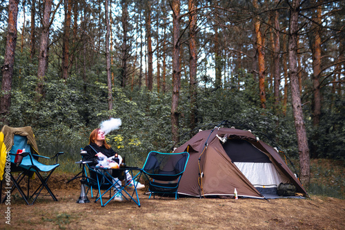Obraz na płótnie A woman exhales smoke have hookah, sits near folding table alone, drink drinks, have satisfied expressions. Pine forest, summer day. Travel and vacation. Camping tent. w salonie