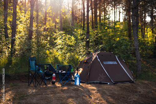Selective focus. Tourist camp. Brown tent and folding chairs with table. Camping equipment. Pine forest. Sunny summer day. Travel and active lifestyle. No body. Hookah flask, water bottle