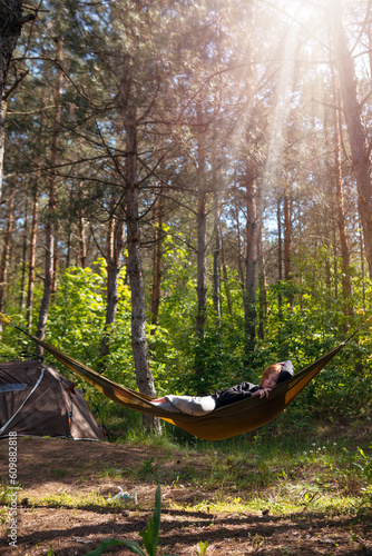 Woman relaxing in the hammock in the middle of a pine forest, watching sundown. Slow life concept. Hipster. Camping tent. Vertical photo