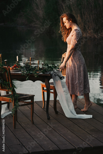 Woman on sunset on dreamy picnic by the water. Romantic evening portrait of female at her 30s. Person on celebration on festive decorated location on pier. Flowers and candles on table photo